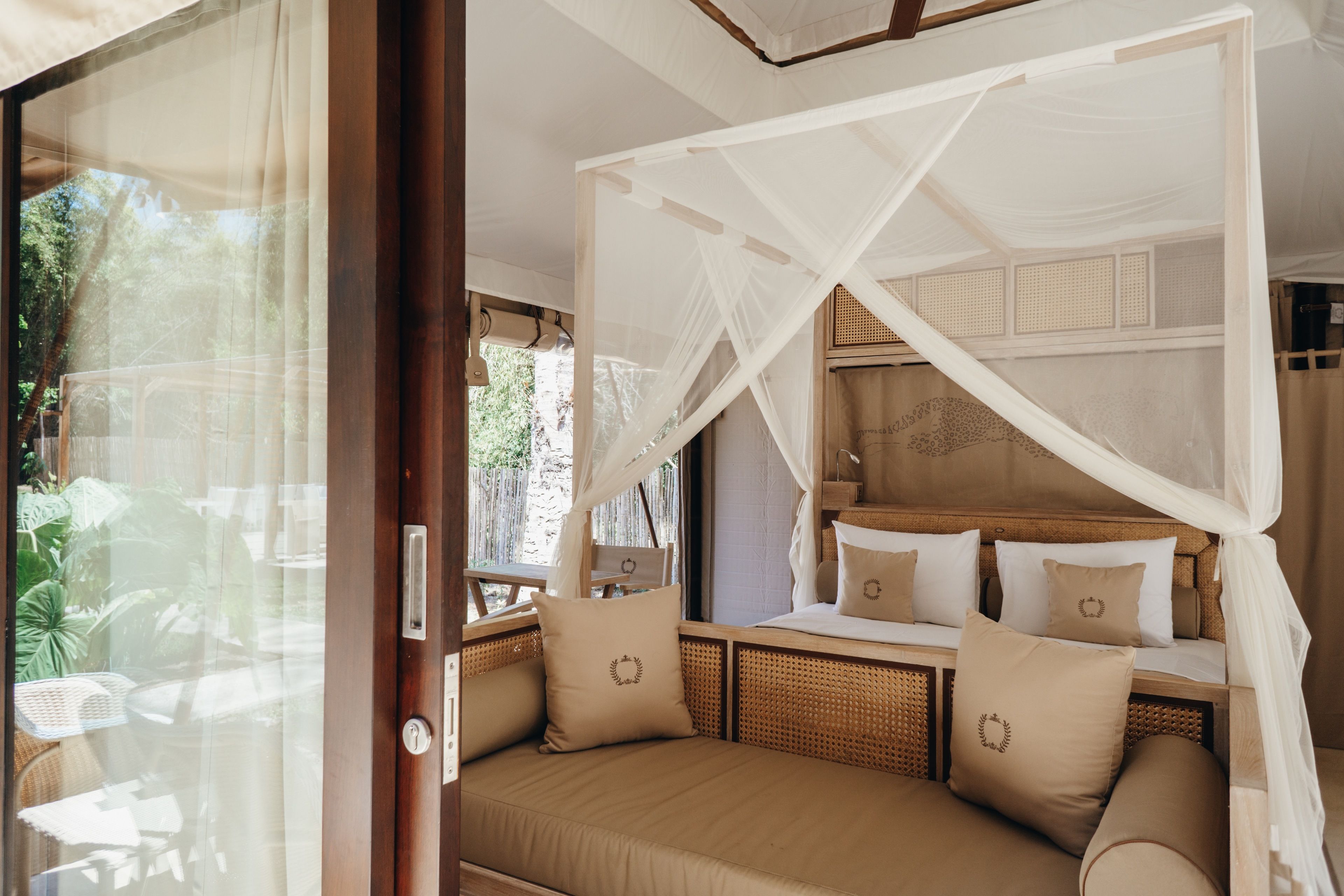A room with a bed under a canopy, a window with curtains, and a view of greenery outside.