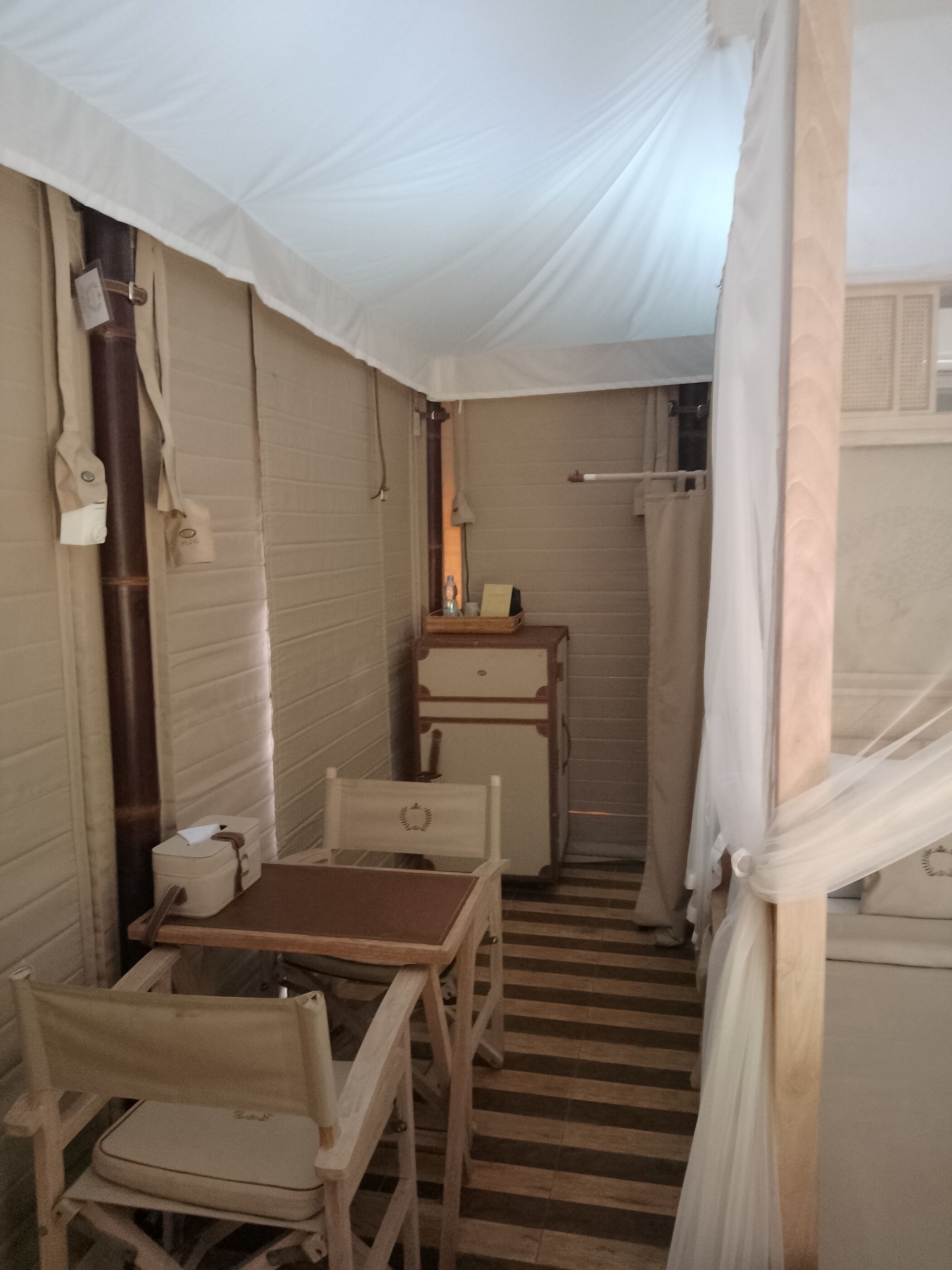 A compact, enclosed room with a bed, a small table, and a chair. There is a wooden bench, a suitcase, and a small cabinet.
