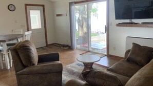 Ein Wohnzimmer mit einer Couch, Sesseln, einem Couchtisch, einem Fernseher und einer Glasschiebetür, die zu einem Außenbereich führt.