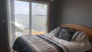 Ein Schlafzimmer mit einem großen Bett, Blick auf ein Gewässer durch eine Glastür und einem Holz-Kopfteil.