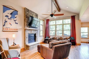 A living room with a stone fireplace, a television mounted on the wall, a ceiling fan, and large windows with red curtains.