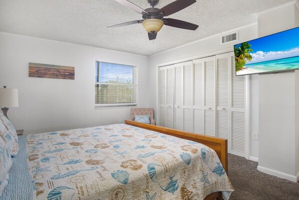 A bedroom with a bed, a ceiling fan, a window, a closet, and a TV displaying a beach scene.