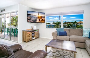 A living room with a sofa, coffee table, TV, wooden cabinet, and a view of the ocean through large windows.