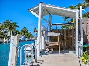 A wooden deck with a metal railing, a covered seating area, and a view of a marina with boats and palm trees.