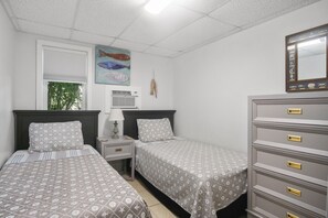 A bedroom with two beds, a dresser, a nightstand, a lamp, a wall-mounted air conditioner, and a fish painting.