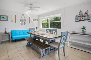 A dining area with a wooden table, blue chairs, a blue sofa, a ceiling fan, and a window with a view of greenery.