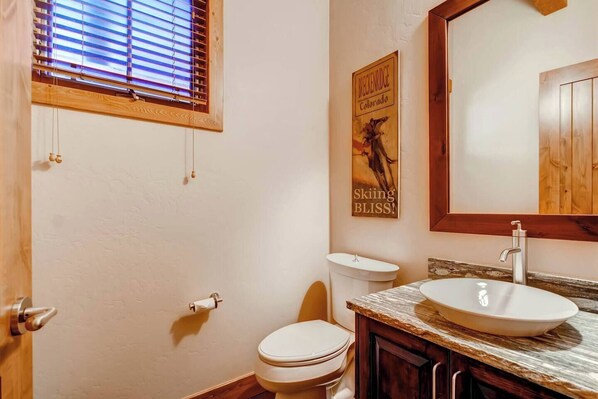 A bathroom with a toilet, sink, wooden vanity, and a window with blinds.