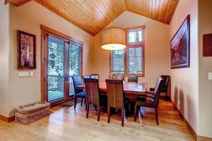 Un coin repas avec une table et des chaises en bois, de grandes fenêtres équipées de stores, un luminaire suspendu et des œuvres d’art encadrées accrochées aux murs.