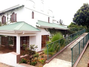 Um edifício branco com telhado verde e um espaço de varanda coberto. Há uma escadaria que leva ao edifício e um guarda-corpo de metal ao lado.