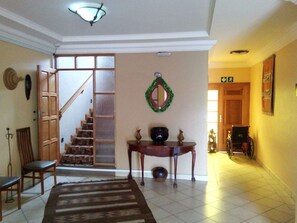 Um corredor com escada de madeira, uma mesa com objetos decorativos e uma cadeira de rodas ao fundo.