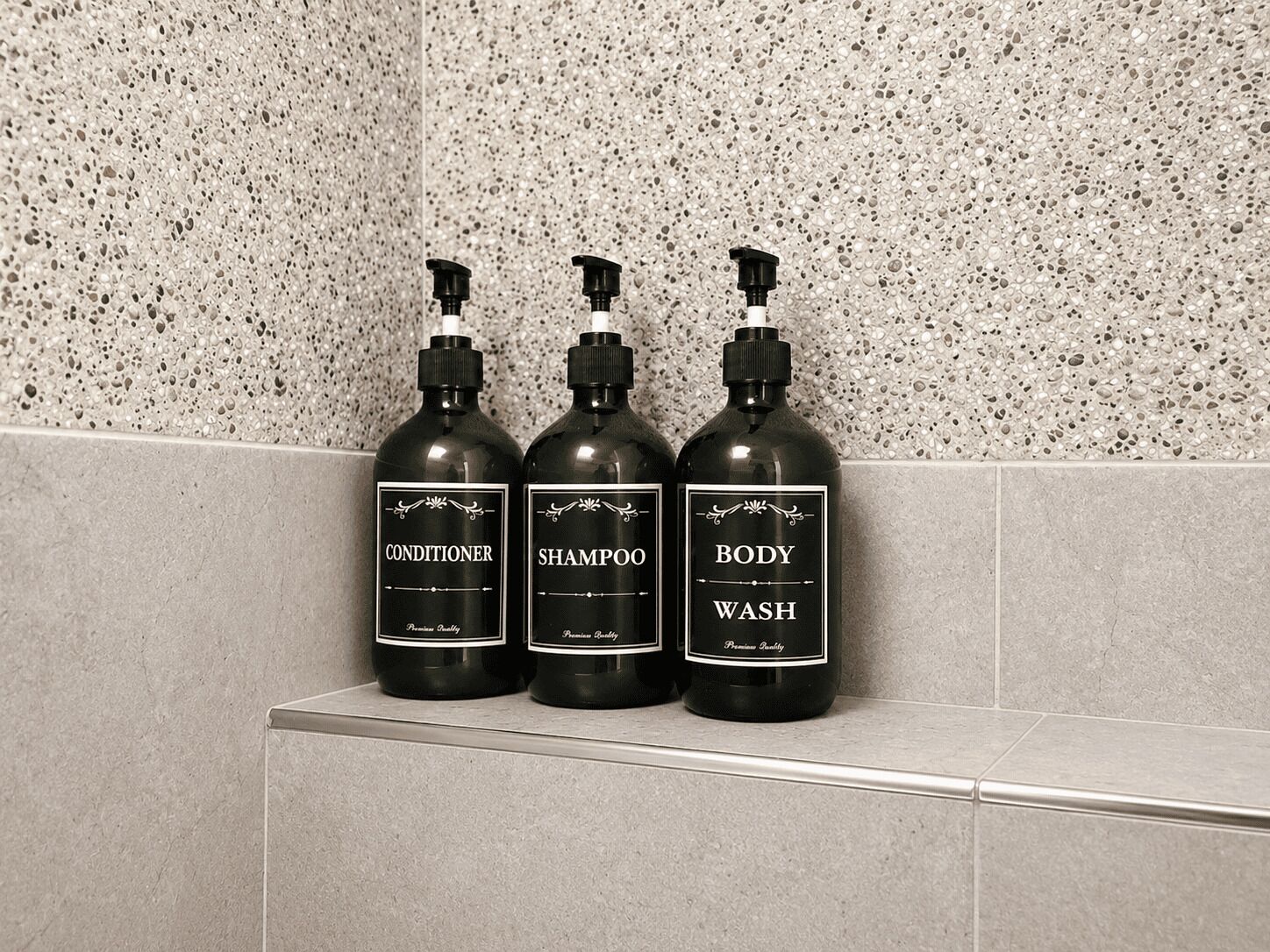 Drie flessen douchegel, shampoo en conditioner op een plank tegen een muren met spikkels.