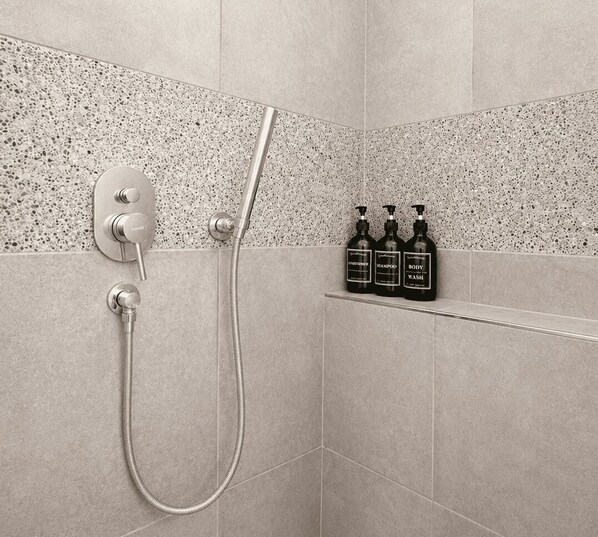 Une douche équipée d’un pommeau de douche à main et d’un panneau de commande fixé au mur. Trois flacons de gel douche sont posés sur une étagère au-dessus de la douche.