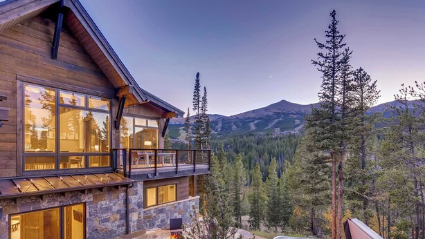 Una cabaña de madera con cimientos de piedra, ventanas amplias, una terraza y un jacuzzi.