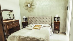 Een slaapkamer met een gestoffeerd hoofdeinde, een grote spiegel, twee nachtkastjes met lampen, een bed met een gewatteerde spreideken en een stoel.