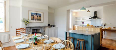 Un coin repas avec une table en bois dressée pour le repas, une cuisine avec un îlot central bleu et une cheminée.