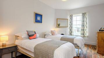 A bedroom with two beds, a nightstand, a dresser, a window with curtains, a painting, and a lamp.