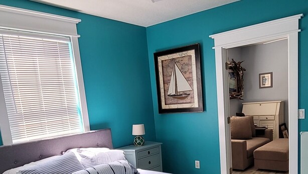 Une chambre à coucher comprenant un lit, un ventilateur de plafond, une table de chevet, une lampe, un tableau encadré et une vue sur une pièce adjacente.