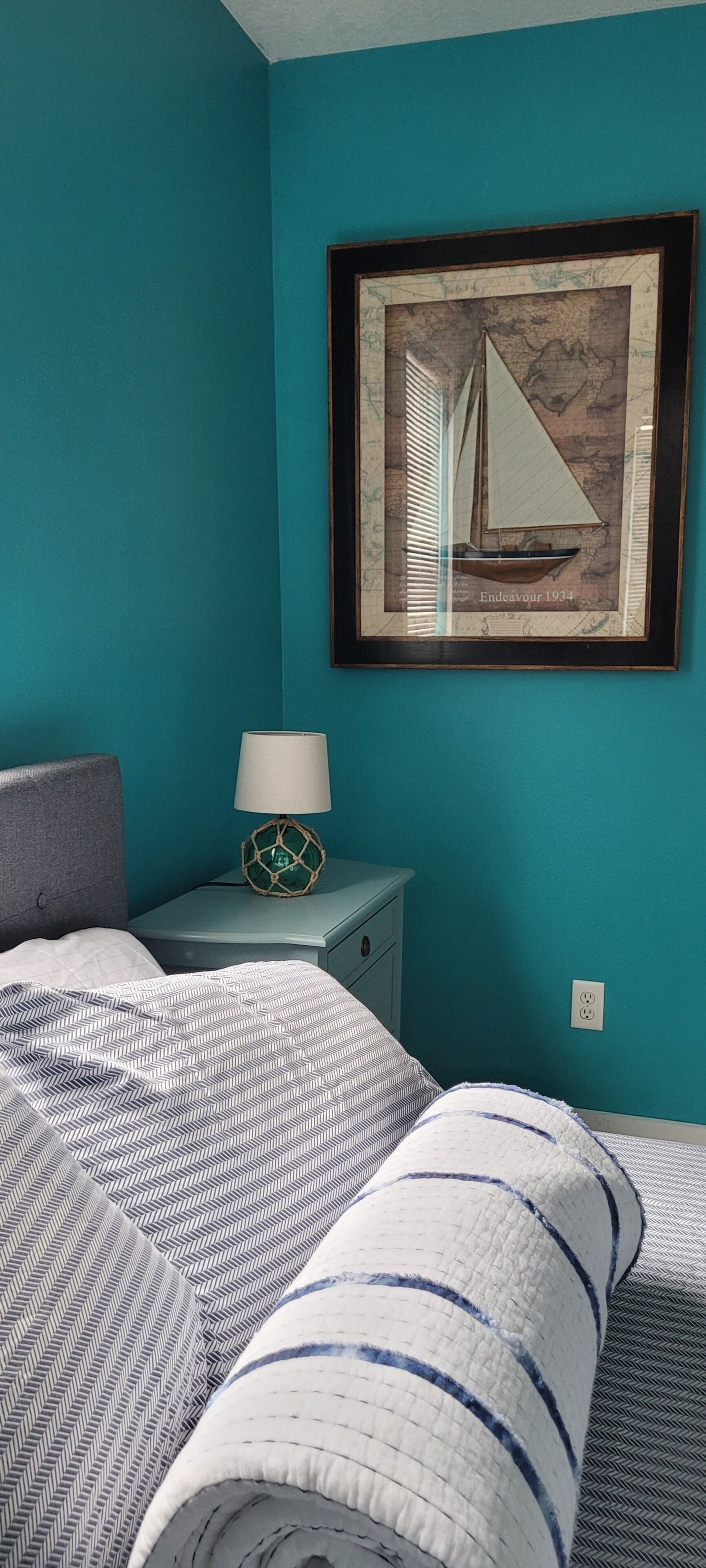 A bedroom with teal walls, a bed with patterned bedding, a nightstand with a lamp, and a framed map on the wall.