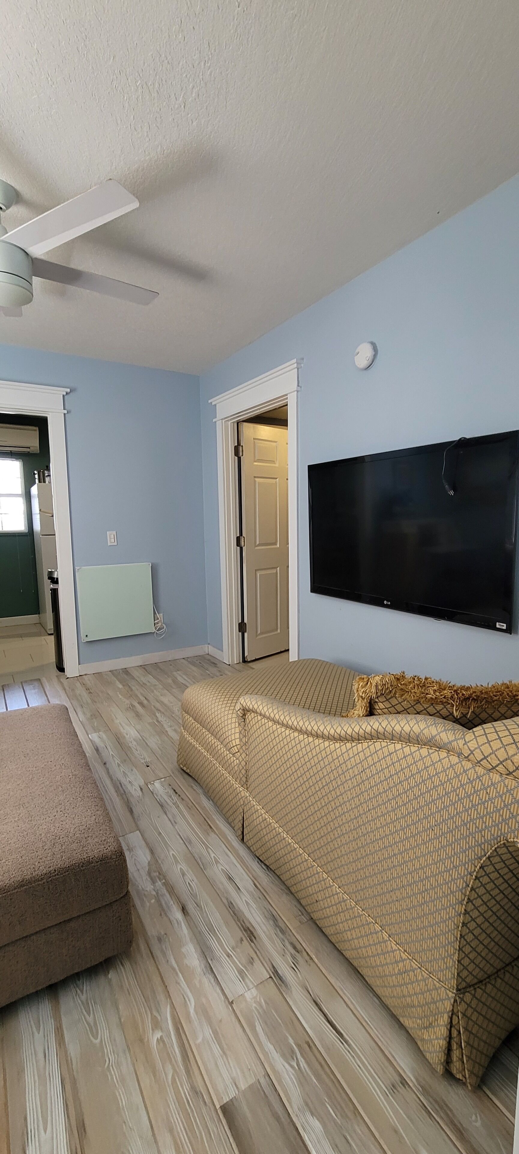 A room with a ceiling fan, a flat-screen TV mounted on the wall, a beige sofa, and a wooden floor.