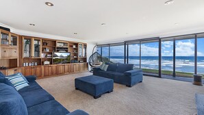 A modern living room with a large TV, wooden shelving, and a view of the ocean.