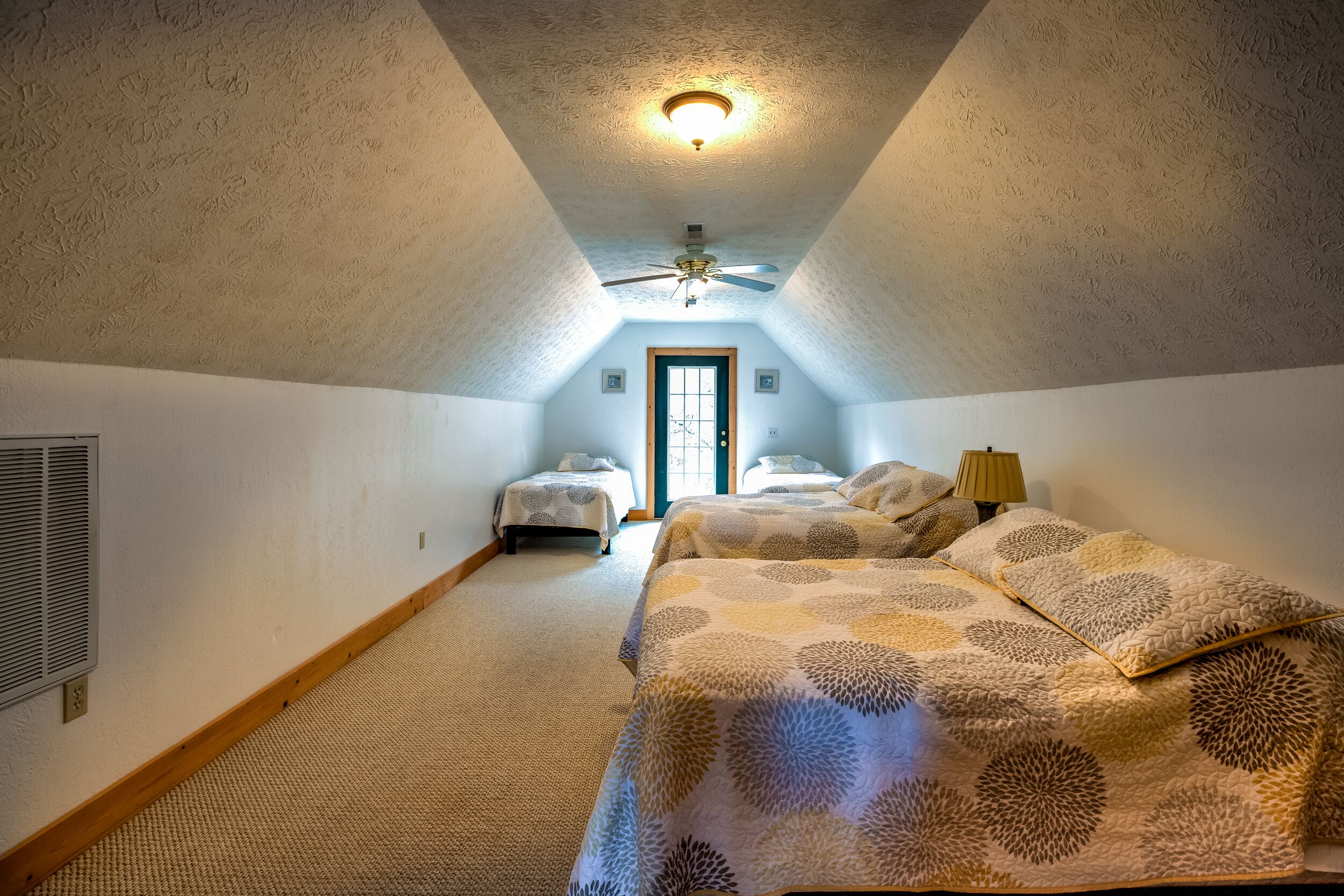 A bedroom with a sloped ceiling, a ceiling fan, a bed with a patterned bedspread, and a nightstand with a lamp.