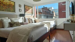 A modern bedroom with a large bed, a bedside table, a lamp, and a view of buildings through a window. - Apartment 3Beds + QueenBed + Balcony + Parking - 6thFLOOR (Quito)