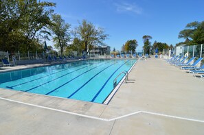 Una piscina con agua cristalina azul, rodeada de sillas de descanso y sombrillas, con árboles y un edificio al fondo.