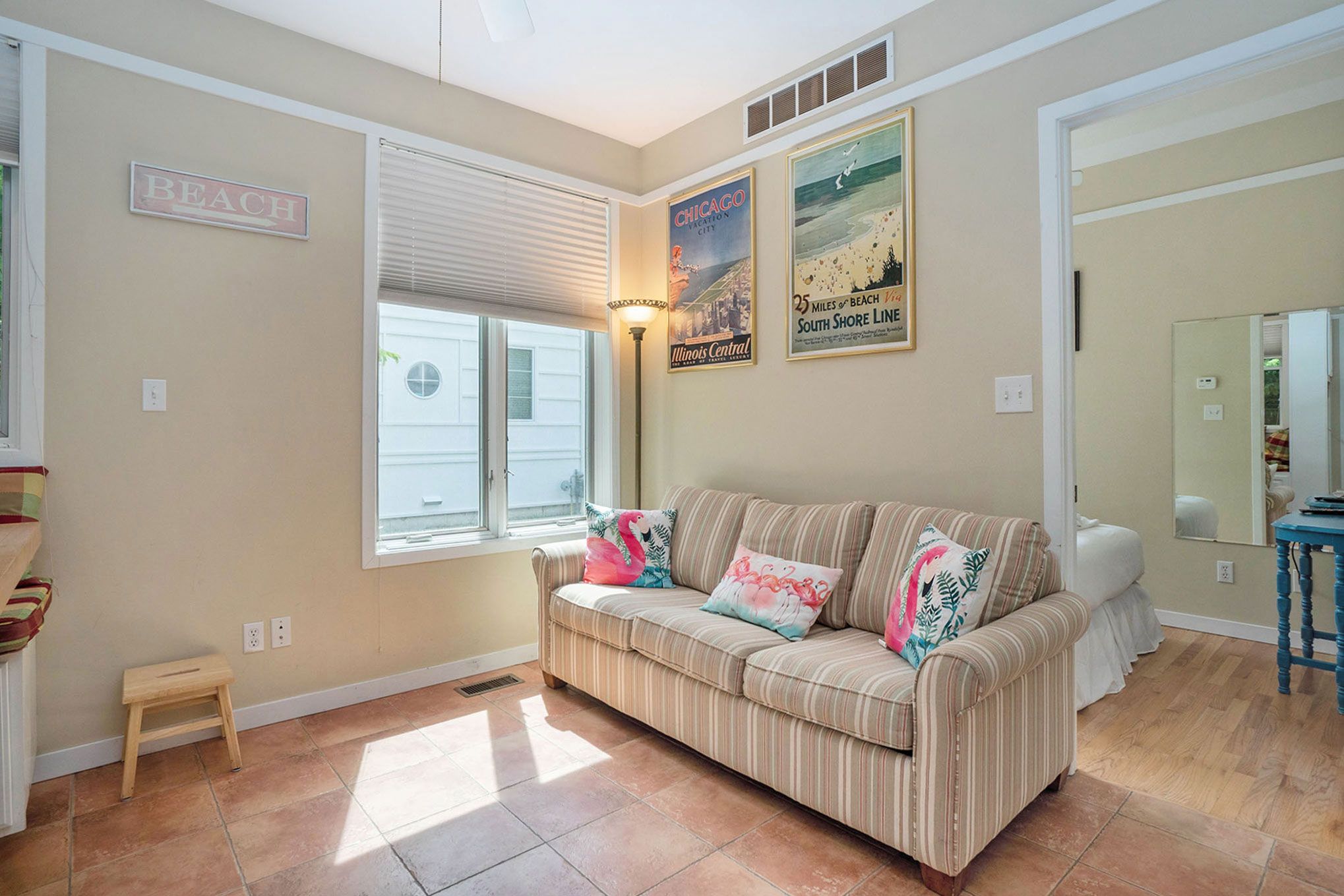 A living room with a striped sofa, a small wooden stool, and a window with blinds. There are two wall-mounted posters and a sign on the wall.