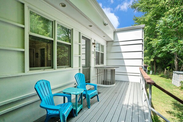 A covered porch with two blue Adirondack chairs and a small table, a white building with large windows, and a grassy yard with trees.