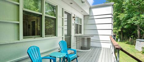 A covered porch with two blue Adirondack chairs and a small table, a white building with large windows, and a grassy yard with trees.