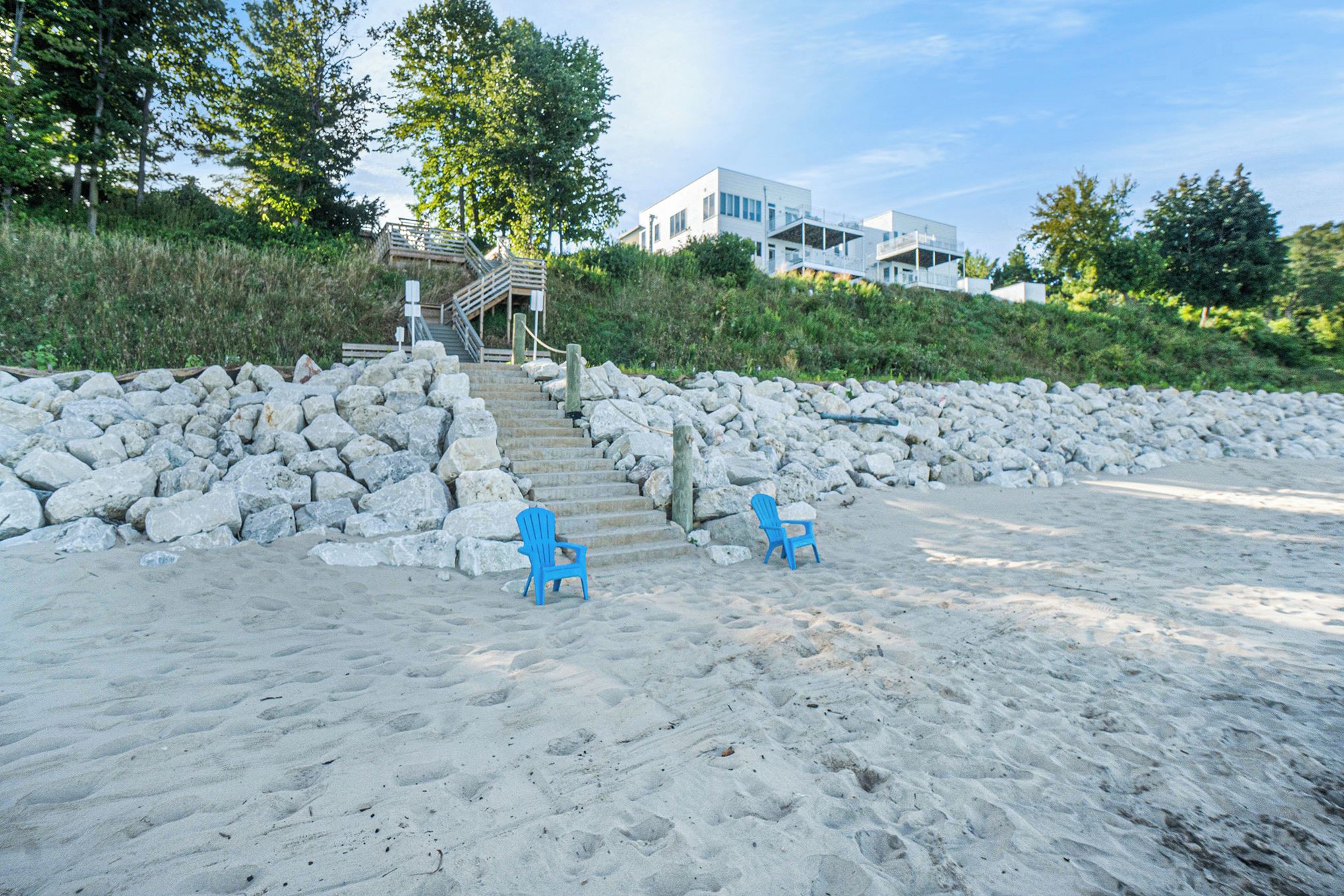 A beach with two blue chairs, a stone wall, and a staircase leading to a building.