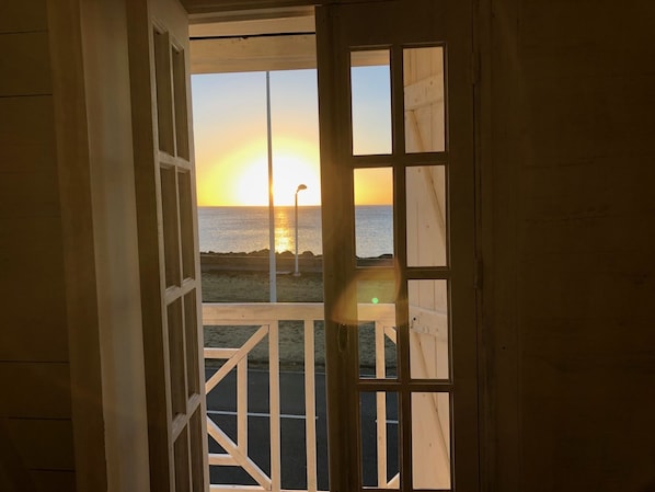 A view through a wooden door with a grid pattern, leading to a balcony overlooking a sunset over the ocean.