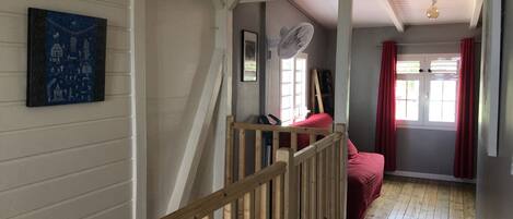 A room with a wooden staircase, a bed with red bedding, a fan, and a window with red curtains.