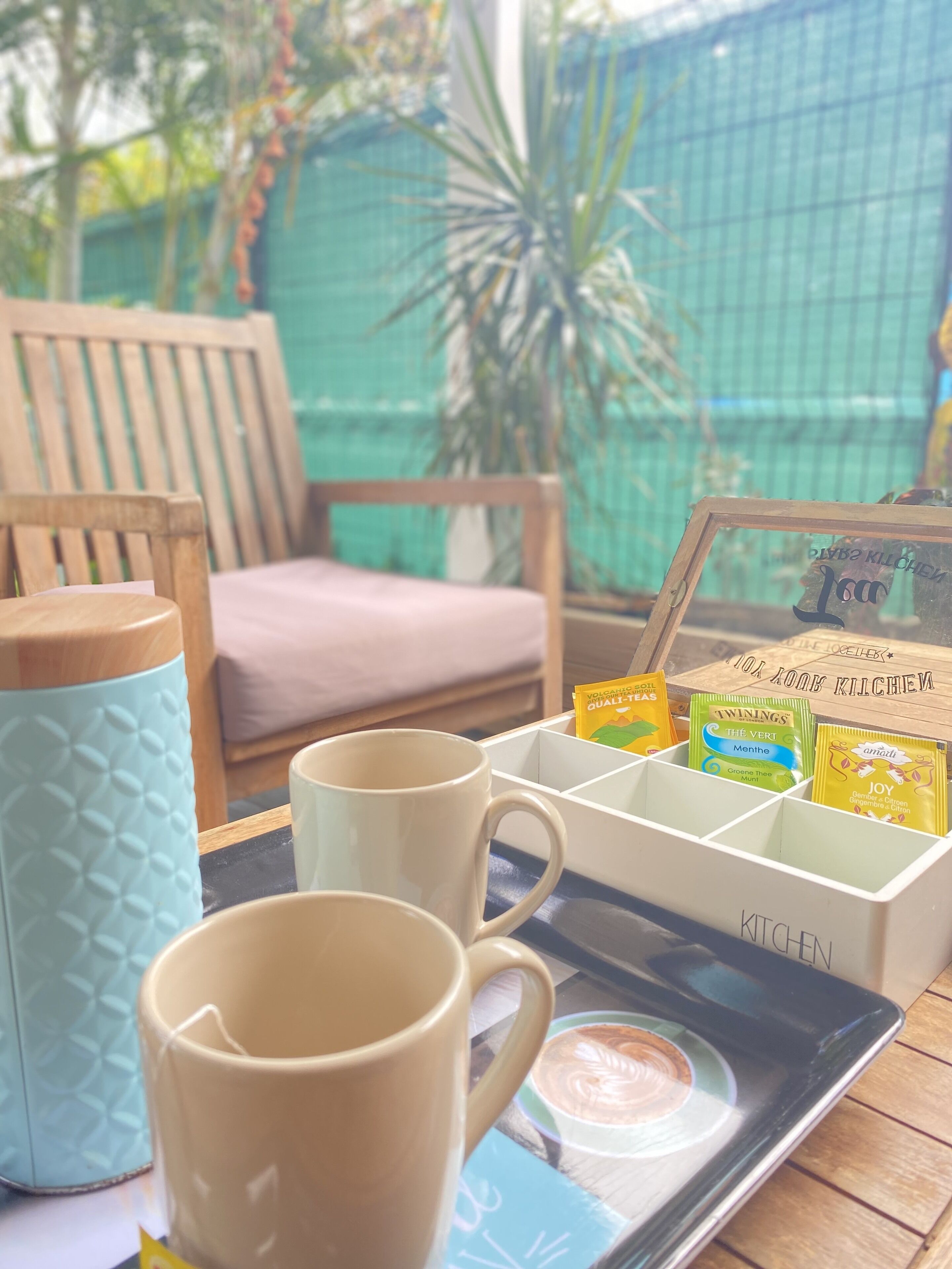 A tray with coffee cups and tea packets on a table, a wooden chair, and a view of a green fence and plants.