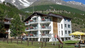 Um hotel com várias andares, varandas, parquinho infantil e montanhas ao fundo.