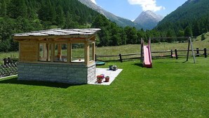 Uma pequena cabana de madeira com base de pedra, uma mesa de piquenique com plantas em vasos, um escorrega e uma cerca de madeira em um campo gramado com montanhas ao fundo.