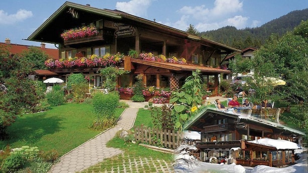 A traditional wooden building with a sloped roof, flower boxes on the balconies, and a paved pathway leading to the entrance. - (2) Ferienwohnung 35 qm mit Balkon (Reit im Winkl)