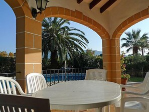 A covered outdoor seating area with a pool view, white chairs, and a table.