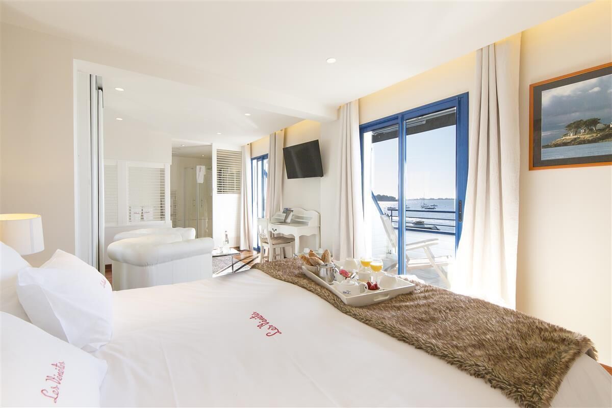 Quarto de hotel com cama, bandeja de café da manhã, televisão e varanda com vista para o mar.