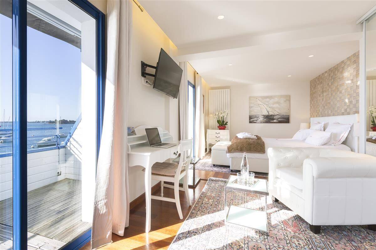 A modern hotel room with a large window, a desk with a laptop, a white sofa, and a bed.