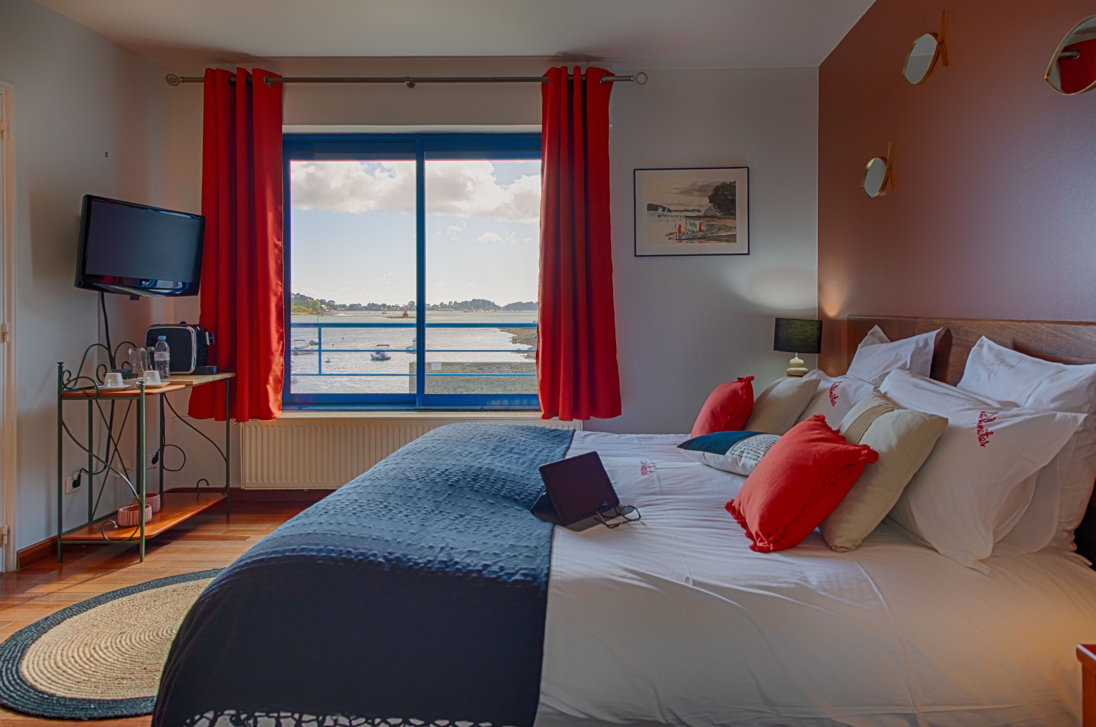 A hotel room with a bed, a desk with a laptop, a television, and a window with red curtains.