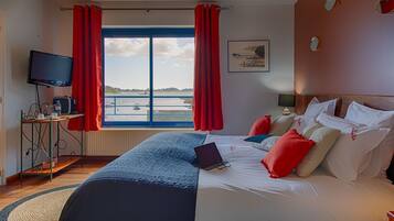 A hotel room with a bed, a desk with a laptop, a television, and a window with red curtains.