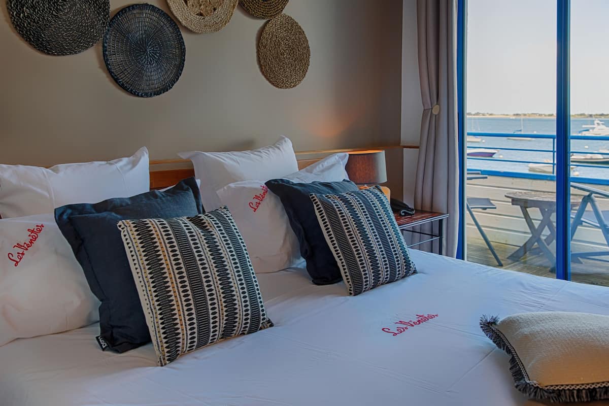 A neatly made bed with multiple pillows, a nightstand with a lamp, and a view of boats through large windows.