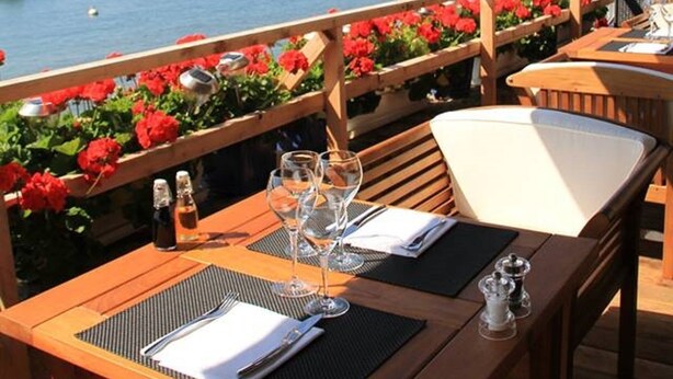 Un área de comedor al aire libre con mesas y sillas de madera, con vista a un puerto deportivo lleno de barcos.