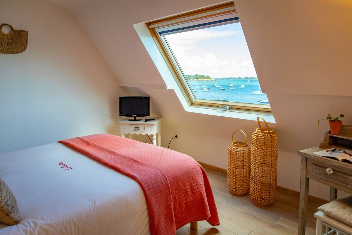 A bedroom with a bed, a television on a small table, a window with a view of boats, and wicker baskets.