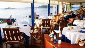 Un restaurante con mesas dispuestas para comer, grandes ventanales con vistas a un cuerpo de agua y gaviotas decorativas sobre las mesas.