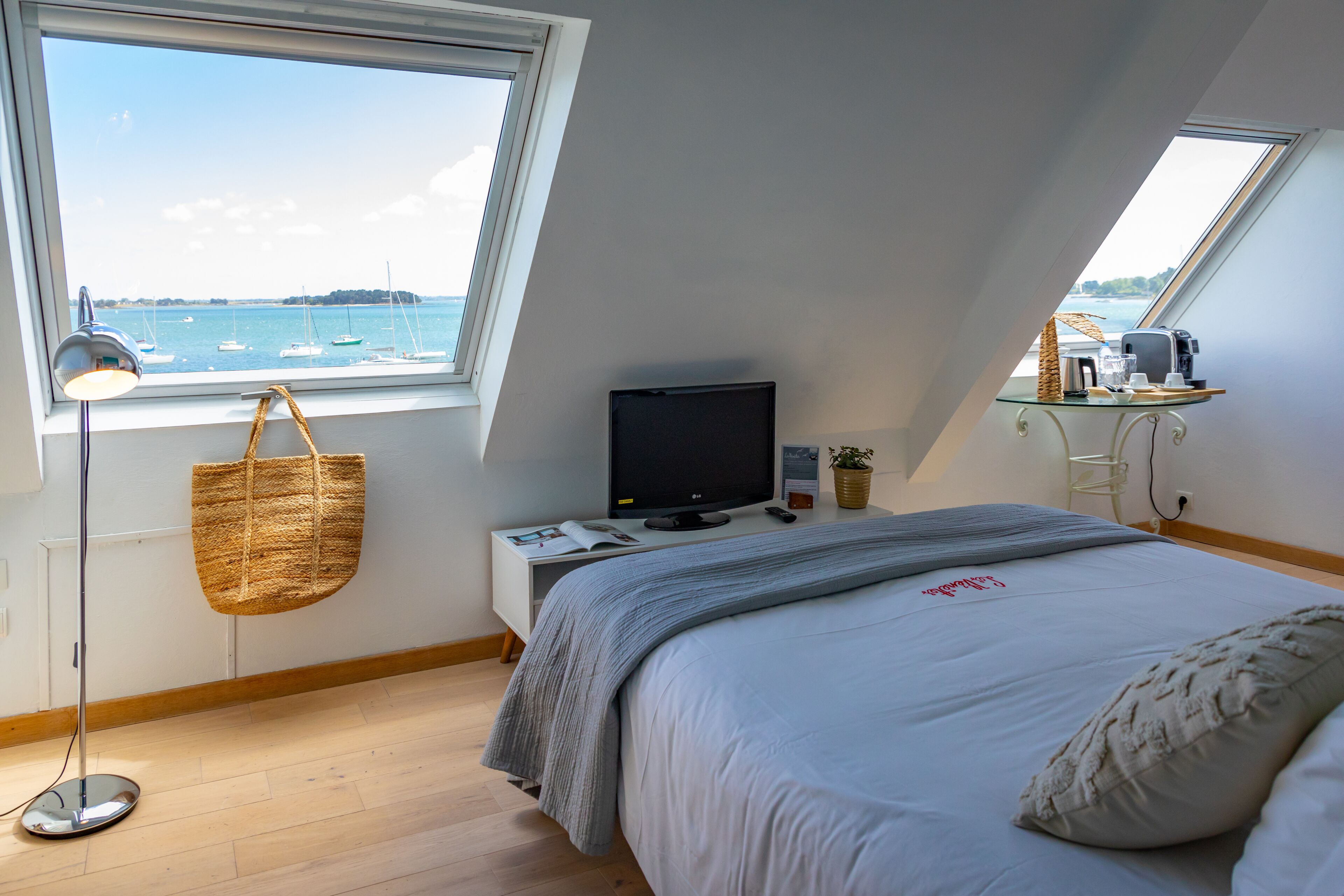 Une chambre avec une grande fenêtre, un lit, une télévision, une table de chevet avec une lampe, et une vue sur les bateaux sur l’eau.