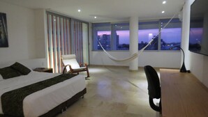 A modern hotel room with a bed, a chair, a desk, and large windows offering a city view.