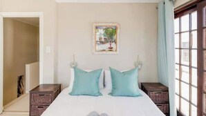 Un dormitorio con una cama, dos almohadas, una mesita de noche, una ventana y una puerta.