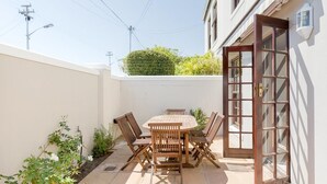Un patio con una mesa y sillas de madera, rodeado de plantas y una pared blanca.